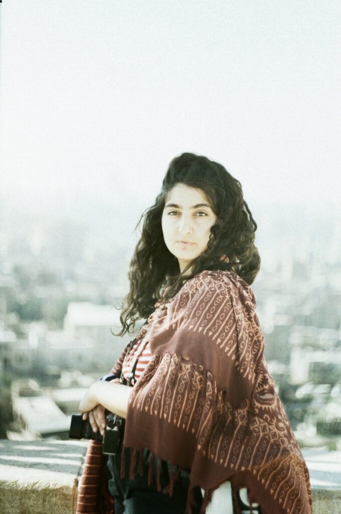 Φωτογραφία πορτραίτο μιας νεαρής κοπέλας με καστανά αφράτα μαλλιά ποζάρει με καφέ πασμίνα μπροστά από κάποια θέα που χάνεται στην ομίχλη. 