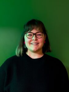 Έγχρωμη φωτογραφία πορτρέτου μιας γυναίκας που στέκεται μπροστά σε ένα ενιαίο πράσινο φόντο και χαμογελάει στην κάμερα. Έχει κοντό σκούρο καρέ με αφέλειες, φορά γυαλιά και φωτεινά ροζ σκουλαρίκια. Φορά μαύρο πουλόβερ, ενώ ο φωτισμός δημιουργεί μια διακριτική πράσινη απόχρωση στο πρόσωπό της και στο φόντο.