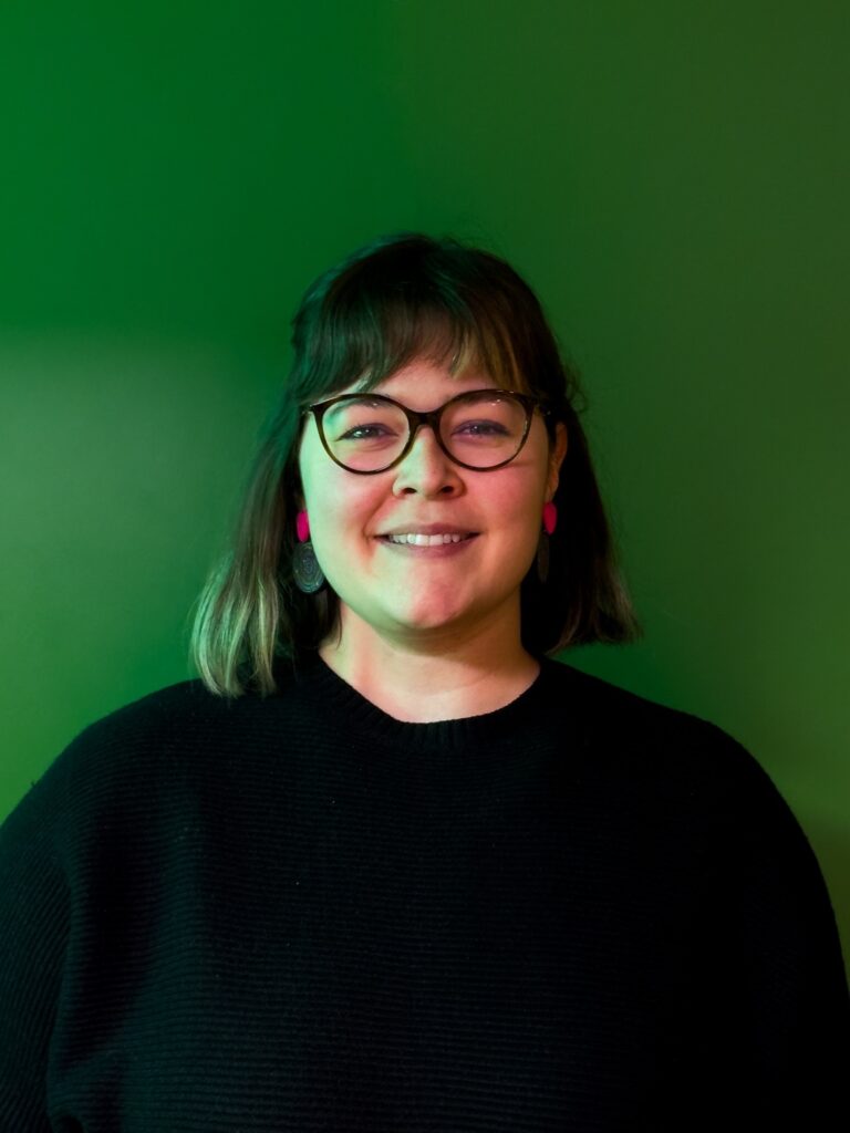 Έγχρωμη φωτογραφία πορτρέτου μιας γυναίκας που στέκεται μπροστά σε ένα ενιαίο πράσινο φόντο και χαμογελάει στην κάμερα. Έχει κοντό σκούρο καρέ με αφέλειες, φορά γυαλιά και φωτεινά ροζ σκουλαρίκια. Φορά μαύρο πουλόβερ, ενώ ο φωτισμός δημιουργεί μια διακριτική πράσινη απόχρωση στο πρόσωπό της και στο φόντο.