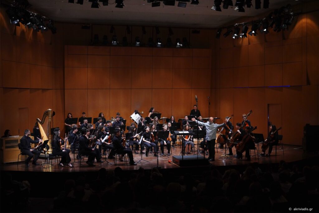 Συναυλία της Underground Youth Orchestra σε αίθουσα του Μέγαρο Μουσικής Αθηνών. Οι μουσικοί κάθονται σε ημικύκλιο επί σκηνής, κρατώντας κυρίως έγχορδα όργανα, με αναλόγια μπροστά τους, ενώ στο πίσω μέρος διακρίνεται μικρή ομάδα χορωδών. Στα αριστερά μια άρπα. Στο κέντρο, πάνω σε βάθρο, ο διευθυντής ορχήστρας στέκεται με ανοιχτά τα χέρια, καθοδηγώντας το σύνολο. Το φόντο αποτελείται από ξύλινα πάνελ σε ζεστούς καφέ τόνους και ψηλά φαίνονται τα φώτα της σκηνής, ενώ στο κάτω μέρος διακρίνεται το κοινό στο σκοτάδι.