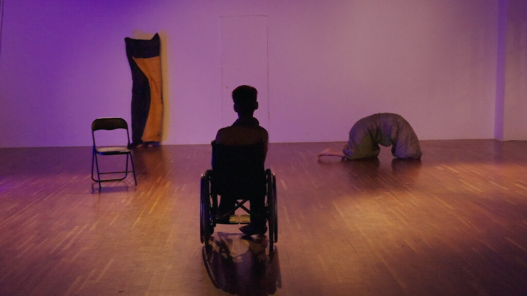 Μια γυναίκα με κοντά μαλλιά σε αμαξίδιο παρακολουθεί δύο άτομα καλυμμένα εξ ολοκλήρου με υπνόσακους να κινούνται σε έναν χώρο με ξύλινο πάτωμα κάτω από ζεστούς φωτισμούς. Το ένα σώμα στηρίζεται στα τέσσερα στο πάτωμα δεξιά, ενώ το άλλο στέκεται όρθιο στον πίσω τοίχο με μια μαύρη καρέκλα λίγα μέτρα μπροστά του ως το μόνο σκηνικό αντικείμενο.