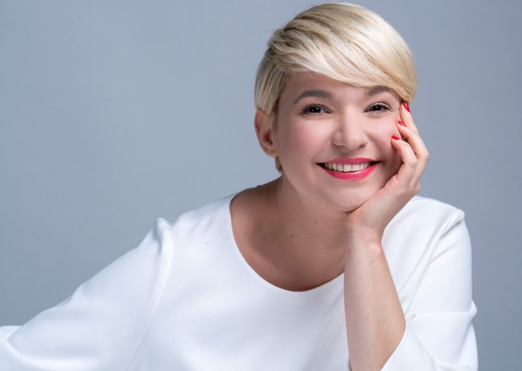 Φωτογραφία πορτραίτου της Νάντιας Κοντογεώργη. Με καστανά μάτια και ξανθά κοντά μαλλιά με χωρίστρα στο πλάι στηρίζει απαλά το χαμογελαστό της πρόσωπο πάνω στην παλάμη του αριστερού της χεριού. Το έντονο ροδοκόκκινο κραγιόν της και τα κόκκινα νύχια της έρχονται σε αντίθεση με την ανοιχτόχρωμη επιδερμίδα της και την ολόλευκή της μπλούζα της.