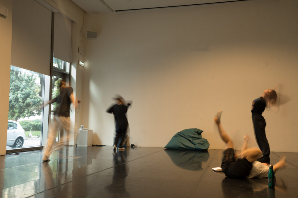 Σε έναν ψηλοτάβανο χώρο με μαύρο πάτωμα χορού, τέσσερα άτομα με άνετα ρούχα βρίσκονται σε κίνηση. Χοροπηδούν, τρέχουν ή κινούν τα μέλη τους στο πάτωμα με ένταση, γεγονός που κάνει τα άκρα τους να θολώνουν στον φακό. Μπουκάλια νερού, τετράδια και άλλα αντικείμενα στον χώρο μαρτυρούν πως ίσως κάνουν το ζέσταμά τους ή την πρόβα τους.