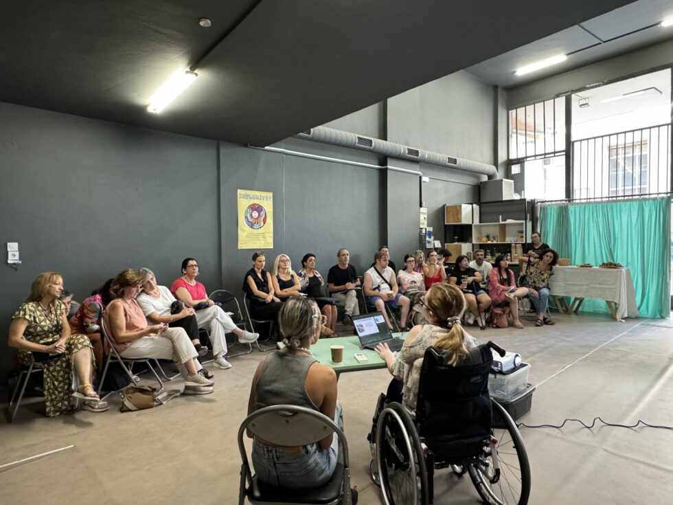 Σε μια απλόχωρη αίθουσα με γκρι πάτωμα και τοίχους, δύο γυναίκες πίσω από ένα βεραμάν τραπέζι με πλάτη σε μας, η μία σε αμαξίδιο και η άλλη καθισμένη σε καρέκλα δίπλα της, με ένα λάπτοπ μπροστά τους συνδεδεμένο σε προτζέκτορα, απευθύνονται σε περίπου 20 άτομα που κάθονται σε ημικύκλιο μπροστά τους. Κάποιοι όρθιοι, κάποιοι καθιστοί παρακολουθούν προσεκτικά αυτό που προβάλλεται σε οθόνη προβολής δίπλα από τις γυναίκες και εκτός του κάδρου της φωτογραφίας.