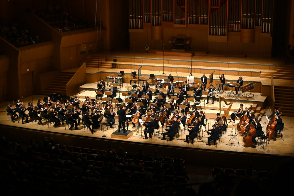 Φωτογραφία μεγάλης συμφωνικής ορχήστρας σε τεράστιο σκηνικό χώρο με επίπεδα και ανοιχτόχρωμο ξύλινο δάπεδο. Τα μέλη της συμφωνικής ορχήστρας φορούν μαύρη επίσημη αμφίεση, κάποιοι παίζουν και άλλοι όχι, αλλά όλοι τους κοιτούν προς τον μαέστρο που διευθύνει μπροστά τους με πλάτη στο κοινό πάνω σε ένα υπερυψωμένο σκαλοπάτι. Μπροστά ξεχωρίζουν τα έγχορδα, στη μέση ένας μικρότερος αριθμός πνευστών και πιο πίσω τα κρουστά. Οι τοίχοι επενδυμένοι κι αυτοί με ξύλο φιλοξενούν στο πίσω μέρος το τεράστιο εκκλησιαστικό όργανο με τους μεταλλικούς αυλούς του σε κάθετη διάταξη.