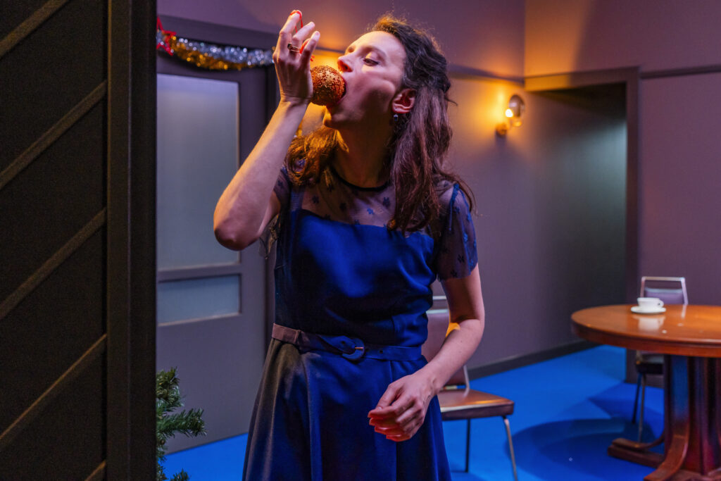 Μια γυναίκα με μπλε φόρεμα με δαντέλα στο πάνω μέρος δαγκώνει λαίμαργα ένα μεγάλο στρογγυλό γλυκό με τρούφα. Από τον διάκοσμο στο σαλόνι γύρω της φαίνεται ότι είναι Χριστούγεννα.