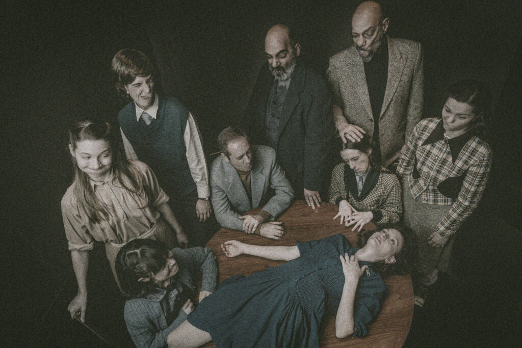 Μια γυναίκα με μπλε φόρεμα βρίσκεται ξαπλωμένη σε ένα ξύλινο στρογγυλό τραπέζι με το χέρι στο στήθος. Το βλέμμα της παγωμένο καρφώνεται ψηλά. Γύρω της, εφτά άτομα ντυμένα με ρούχα που παραπέμπουν στη δεκαετία του '50 την κοιτούν κάνοντας έντονες κοροϊδευτικές γκριμάτσες με τα πρόσωπά τους.