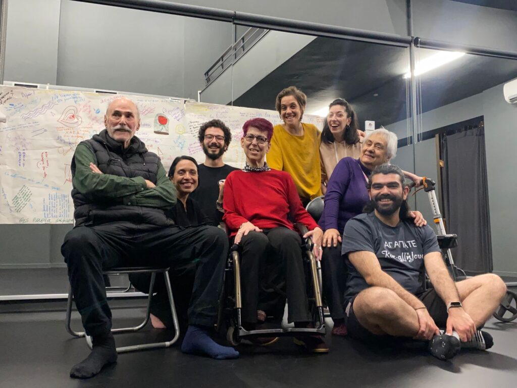 Η συμπεριληπτική θεατροκινητική ομάδα, που αποτελείται από άτομα διαφορετικών ηλικιών και σωματικοτήτων, κοιτά προς την κάμερα χαμογελώντας. Κάποια άτομα κάθονται σε καρέκλες ή στο πάτωμα, άλλα χρησιμοποιούν αμαξίδια και άλλα στέκονται όρθια. Μοιάζει να βρίσκονται σε έναν χώρο πρόβας με καθρέφτες, γκρι τοίχους και γκρι πάτωμα.