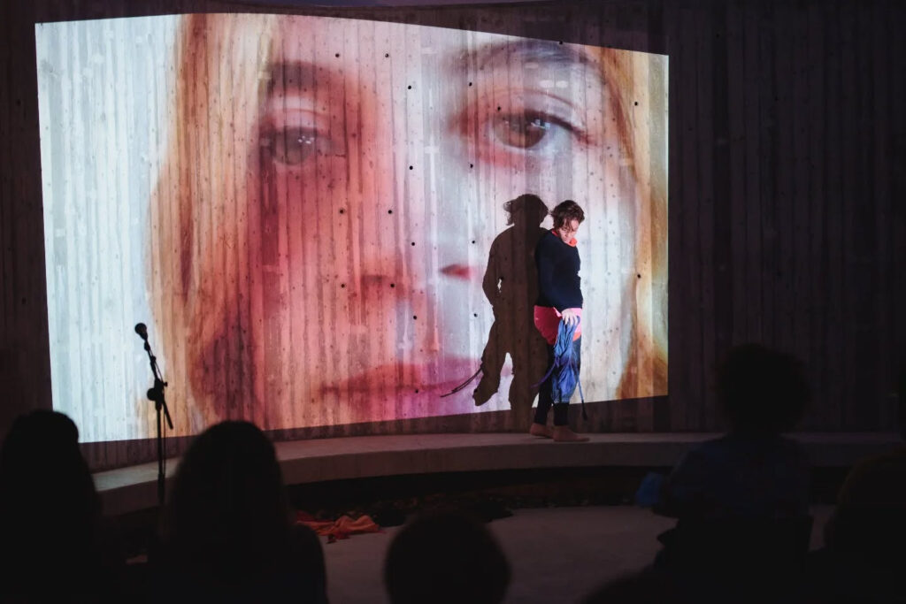 The face of a young blonde woman with brown eyes is projected at a monumental scale onto a corrugated metal sheet, punctured with small holes across its surface. In front of the projection, bathed in its light, a young woman with short brown hair walks barefoot across the stage space, holding in her hand a piece of blue fabric resembling a crumpled shirt or blouse. Looking down, she appears ready to drop it, while other similarly sized fabrics lie scattered across the floor. A microphone on the left side and the heads of audience members in the foreground indicate that this is a moment captured from a live performance.