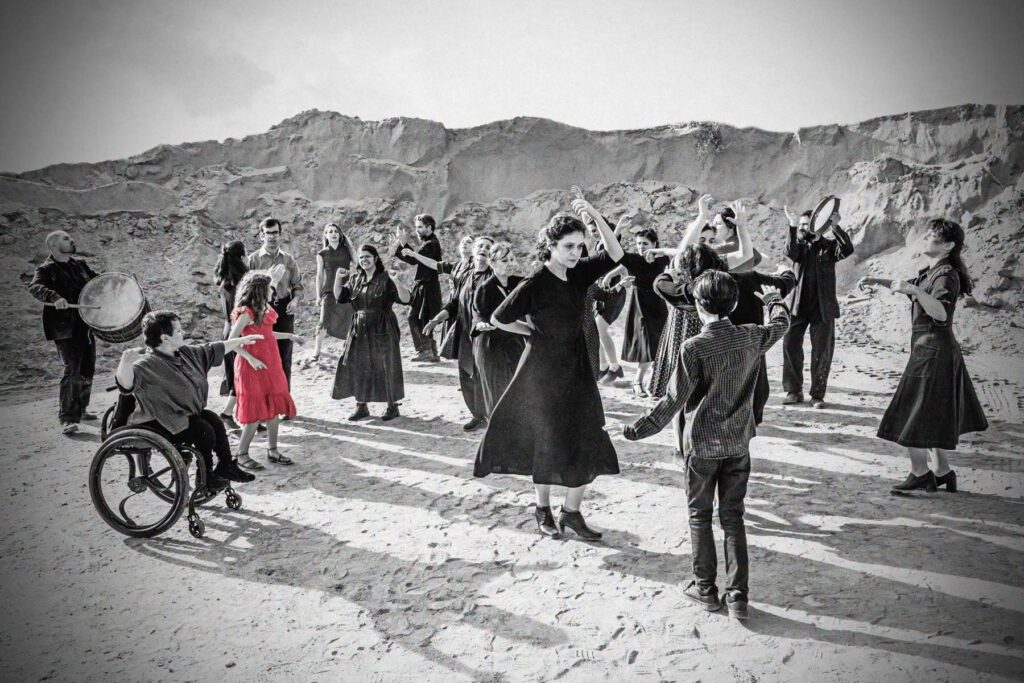 A black-and-white photograph shows a group of people dancing and celebrating in a rocky, barren landscape. Some play tambourines and drums, while others form spontaneous dances. Young and old, children and adults, standing or using wheelchairs, are all dressed in dark clothing — except for a girl of about ten, who wears a red dress.