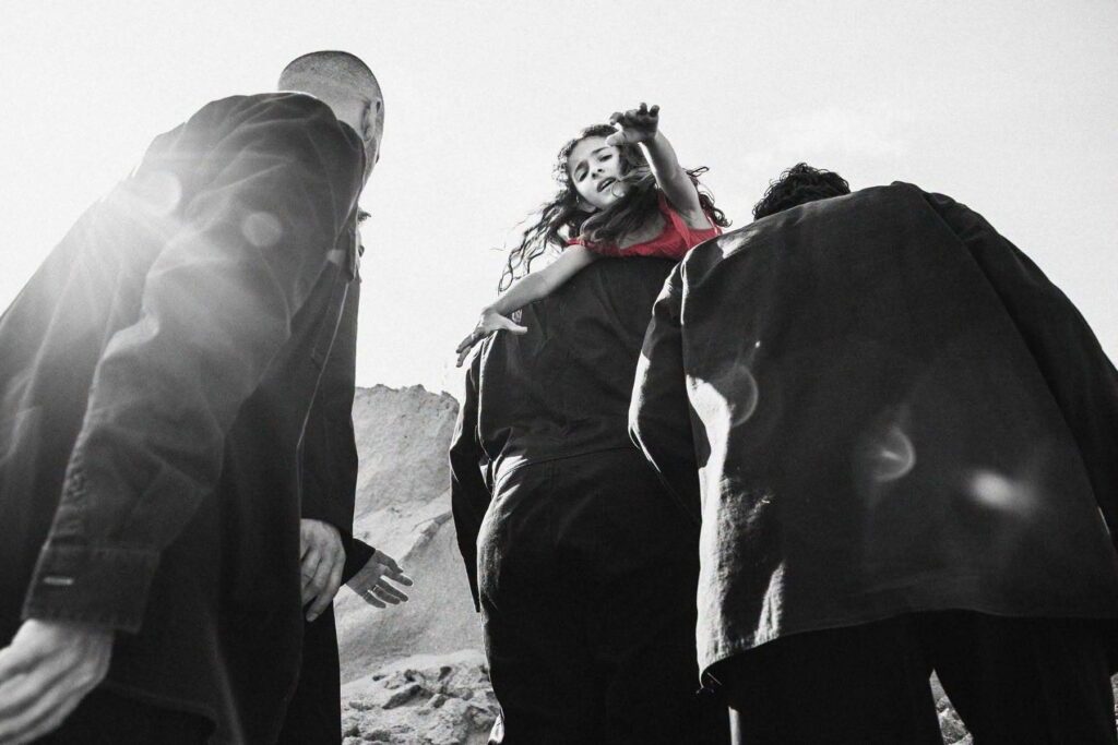 Four men walk among rocks under a bright sun, their backs turned to us. The man in front carries a girl in a red dress in his arms. She turns her face toward us and stretches her hands toward the camera, as if calling for help.