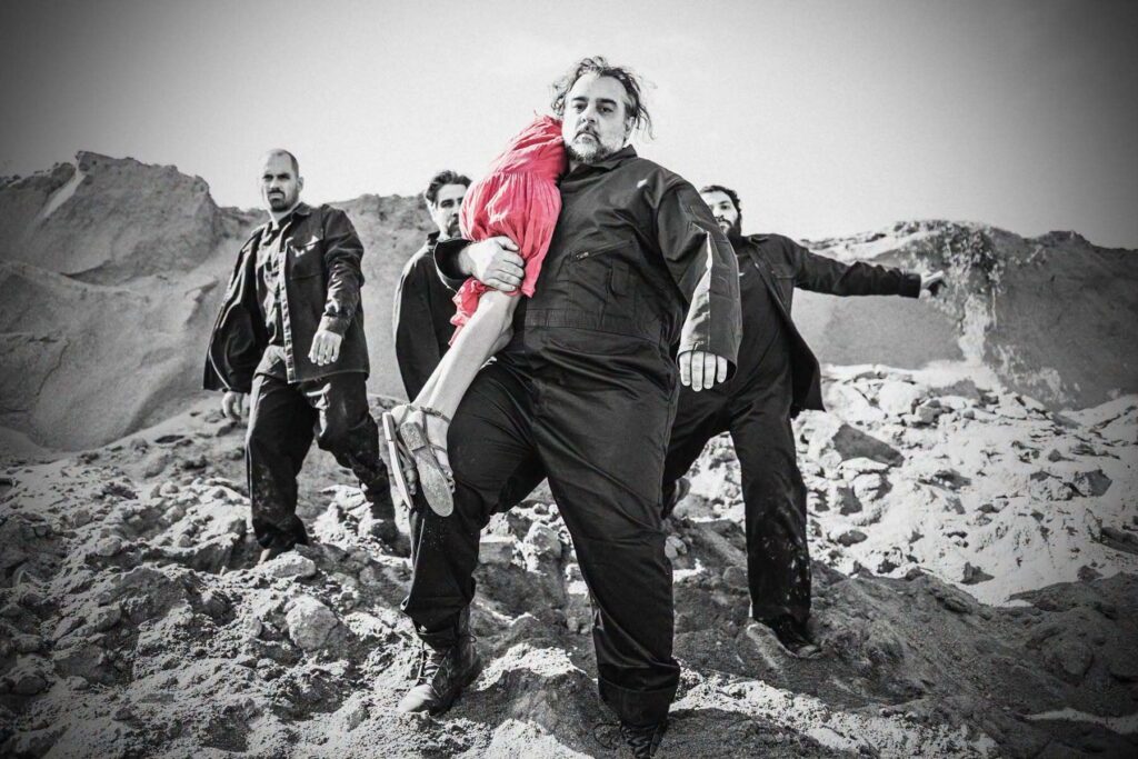 Descending a rocky hill, a man with grey hair and a beard, accompanied by three others behind him, carries the body of a girl in his arms, her torso leaning back over his shoulder. Her red dress is the only colour that stands out in the black-and-white photograph.