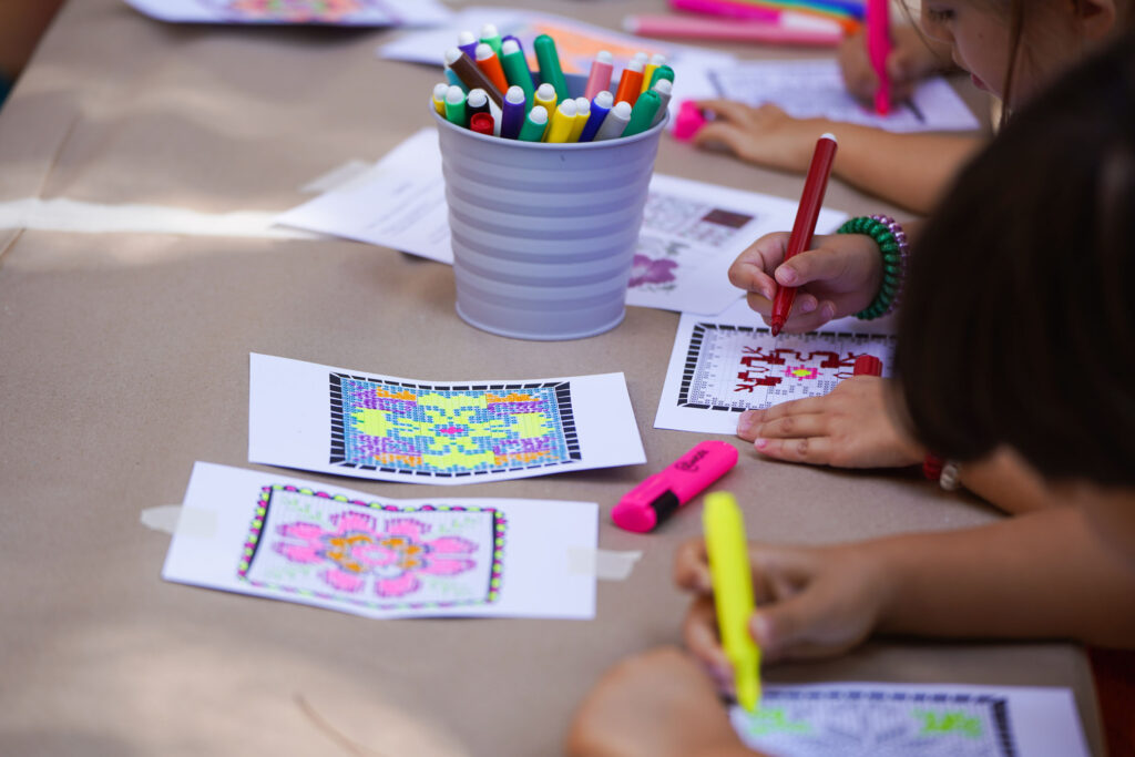 At a table, children use colourful markers to draw images of flowers that appear to be divided into small dots or pixels. Their drawings take on the appearance of mosaics or digital images.