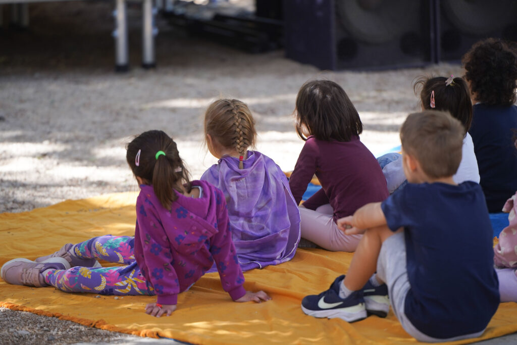 In a sunny outdoor setting, children around the age of eight, wearing colourful clothes, sit on a large orange cloth spread over the gravel. We see them from behind as they appear to be watching something unfolding in front of them, outside the frame of the photograph.