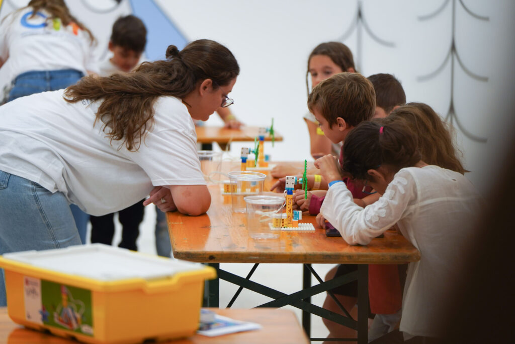 At a wooden table, five children are building something resembling a wind turbine out of LEGO. A woman wearing glasses, a white T-shirt, and jeans leans over the table and looks at their creations with them, as if offering advice or guidance. In the background, other people taking part in the activity can be seen faintly at other tables.