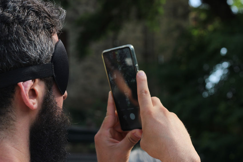 A young bearded man wearing a black sleep mask over his eyes holds his phone upright and uses the camera to capture a lush green tree visible in the distance in front of him.